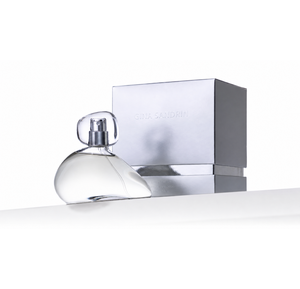 A clear glass perfume bottle with a transparent cap sits in front of a metallic silver box embossed with the brand name 'GINA SANDRINI' on a clean white background.