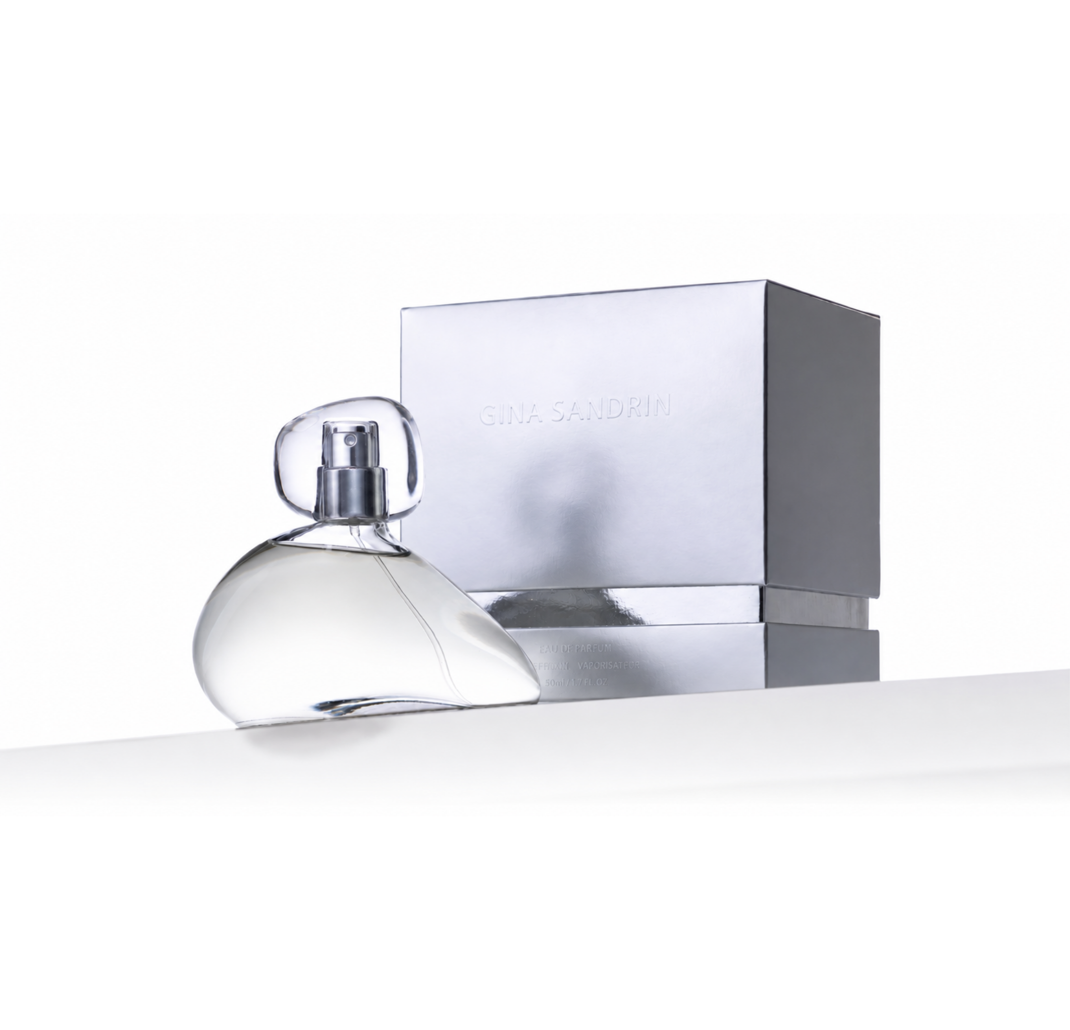 A clear glass perfume bottle with a transparent cap sits in front of a metallic silver box embossed with the brand name 'GINA SANDRINI' on a clean white background.