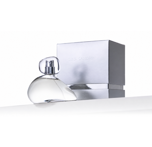 A clear glass perfume bottle with a transparent cap sits in front of a metallic silver box embossed with the brand name 'GINA SANDRINI' on a clean white background.